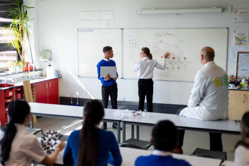 Science classroom with two male teenagers at the front of the class explaining something to the rest of the class. Everybody is listening and looking toward the board.